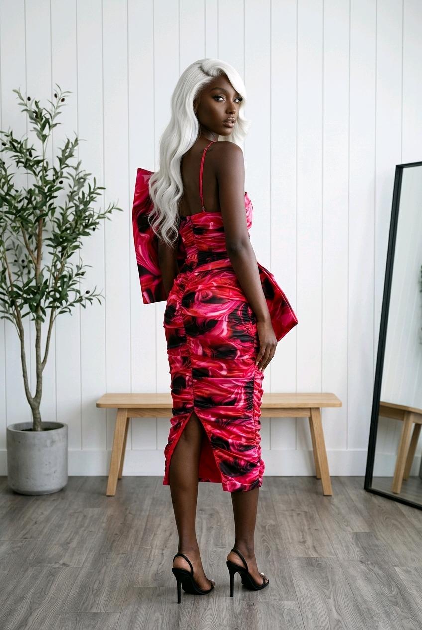 Woman wearing a red abstract print ruched midi dress with oversized sculptural bow detail and fitted silhouette. 
