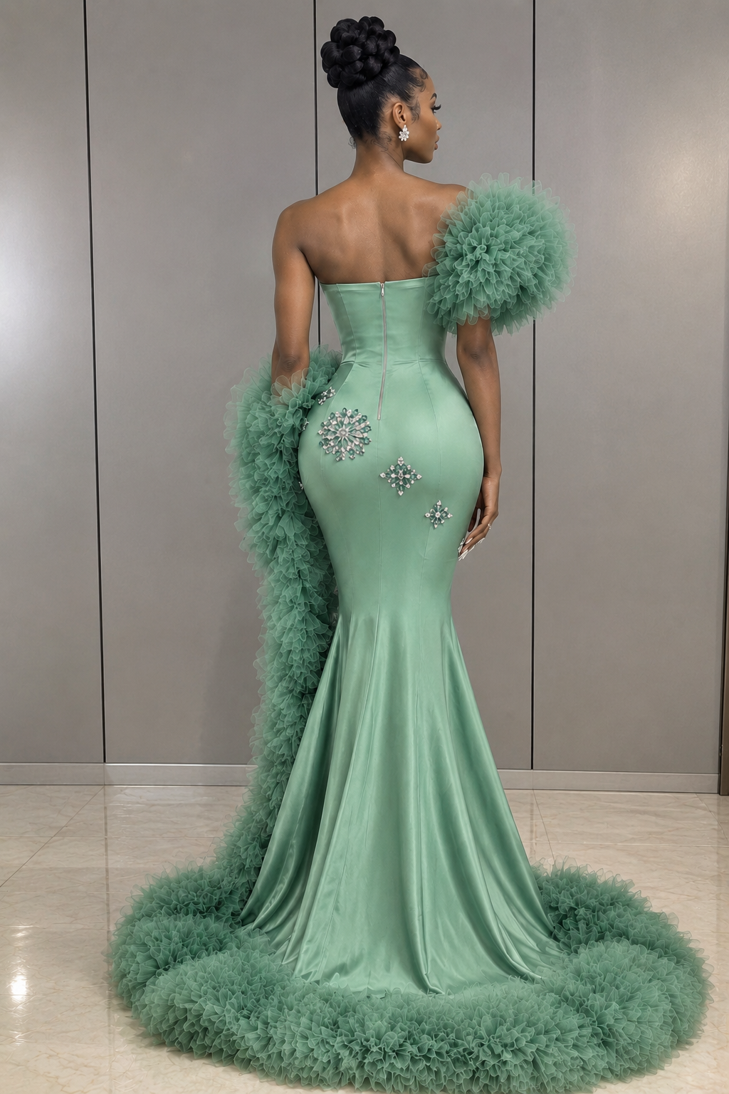 A woman stands with her back to the camera wearing a strapless mint green mermaid gown with voluminous feathered shoulder details, a matching feathered train, and jeweled embellishments on the back, set against a neutral indoor wall.