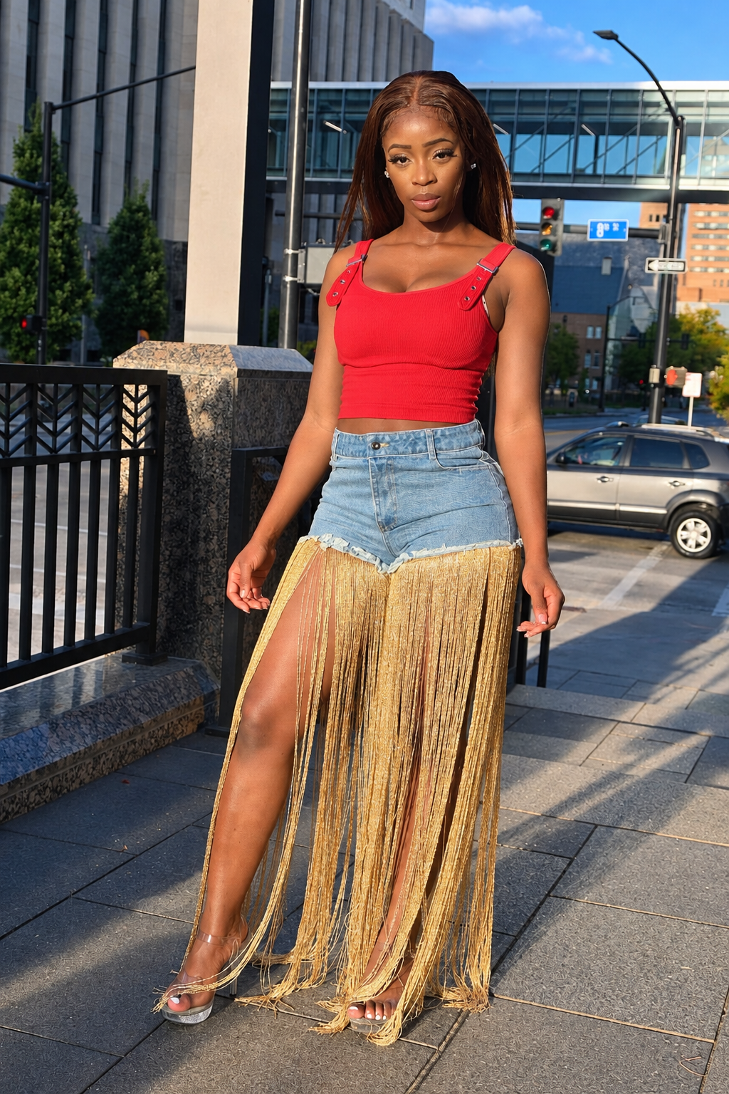 A woman stands on an outdoor city sidewalk wearing a bright red ribbed crop top, light-wash denim shorts, and a long sheer fringe skirt in tan/beige. The background shows buildings, street elements, and a parked car.