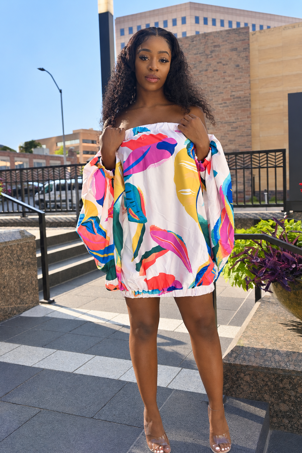 A woman wearing a white off-the-shoulder mini dress with a vibrant multicolor abstract floral print, standing outdoors on stone steps in front of a modern building.