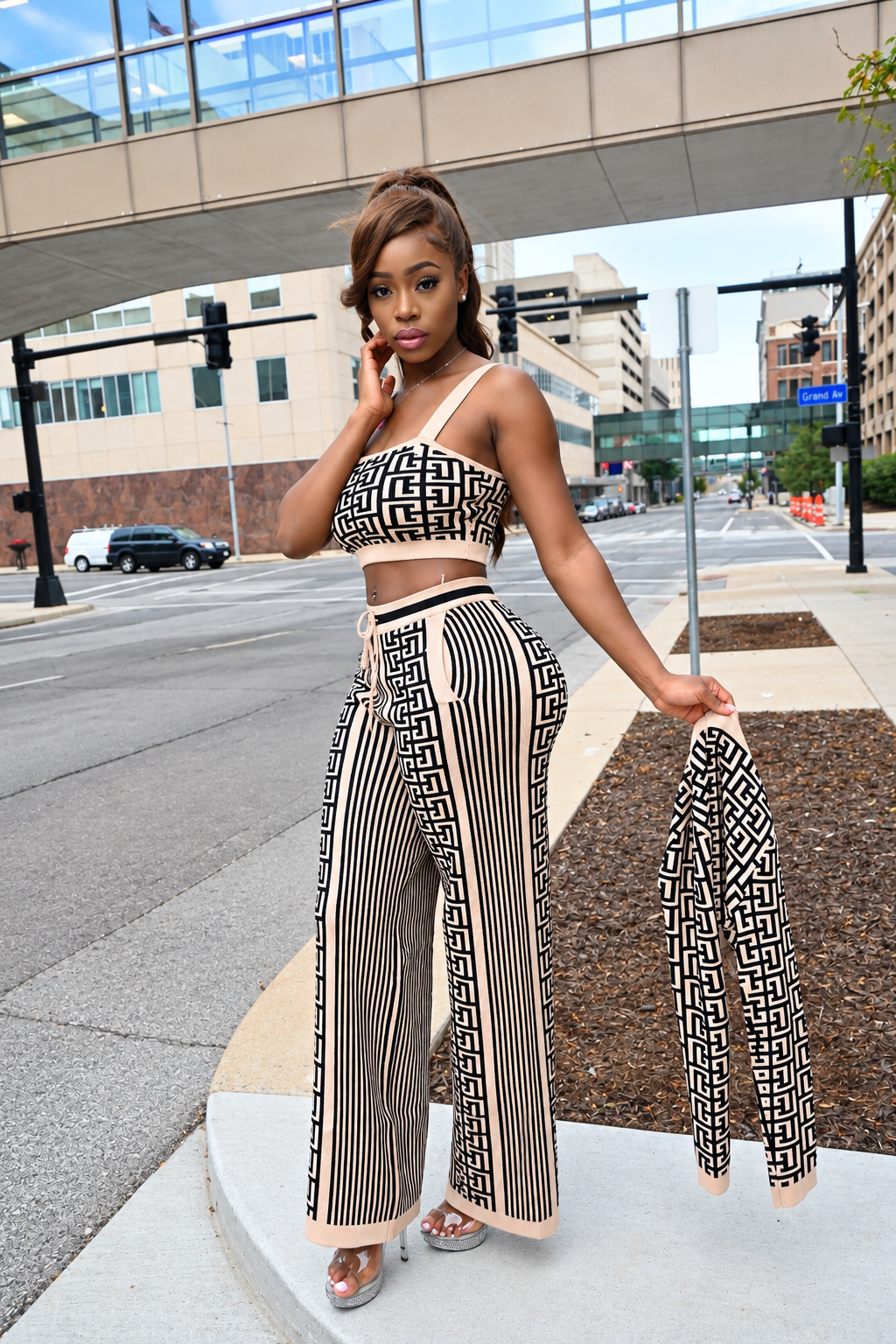 A woman stands on an urban sidewalk wearing a matching two-piece outfit with a black and beige geometric pattern, consisting of a bandeau-style top and wide-leg pants. She holds a matching patterned cardigan or jacket in one hand while posing in front of a city street and bridge.