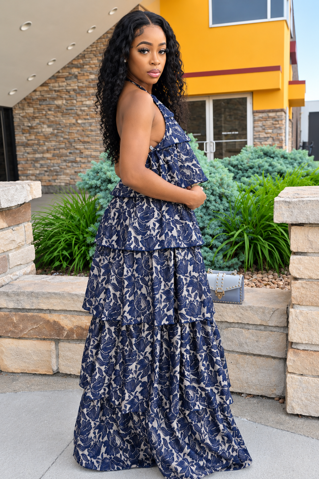 A woman wearing a navy blue and beige floral print tiered halter maxi dress, standing outdoors in front of a modern building with stone walls and green plants.