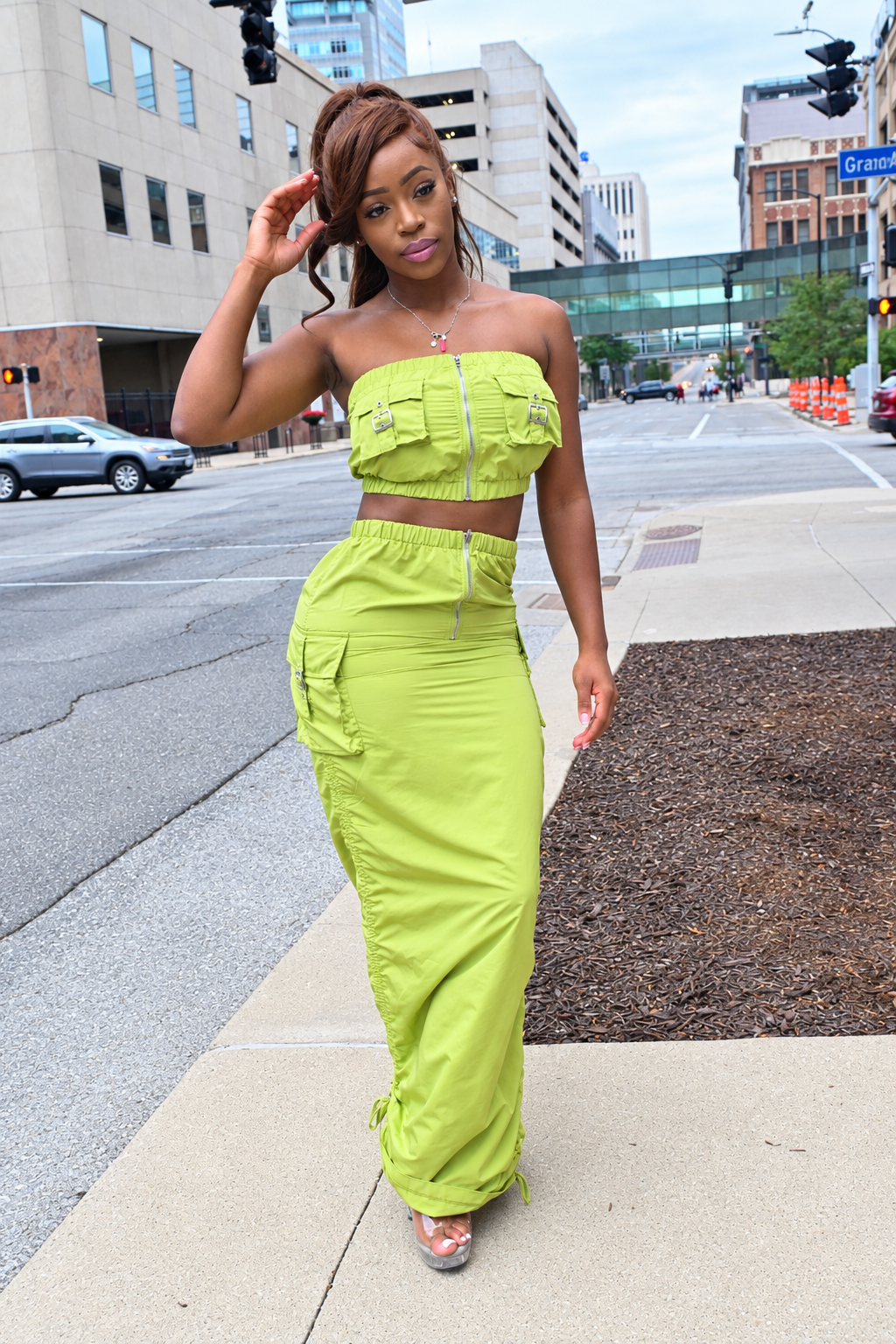 A woman models a bright lime green two-piece outfit consisting of a strapless crop top with a front zipper and matching cargo pants, standing on a city sidewalk.
