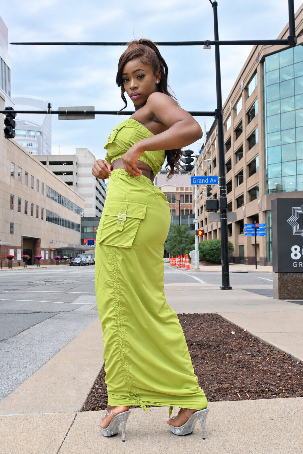A woman posing on an urban sidewalk wearing a lime green two-piece set consisting of a strapless crop top and matching high-waisted cargo maxi skirt, paired with silver high heels.