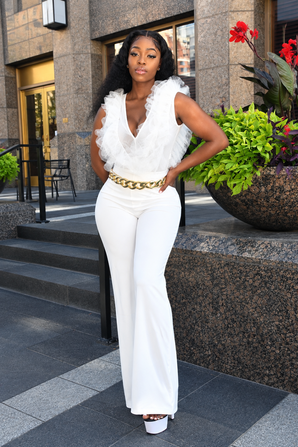 A woman models a white sleeveless jumpsuit with a ruffled tulle bodice and a chunky gold chain belt, standing outdoors in front of a building with stone walls and potted plants.