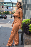 A woman models a matching two-piece satin set in a warm metallic bronze color, consisting of a halter crop top and wide-leg pants, standing on a city sidewalk.