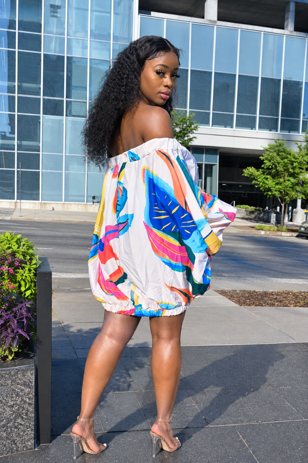 A woman wearing a colorful off-the-shoulder mini dress with a vibrant abstract print, standing outdoors in front of a modern glass building.