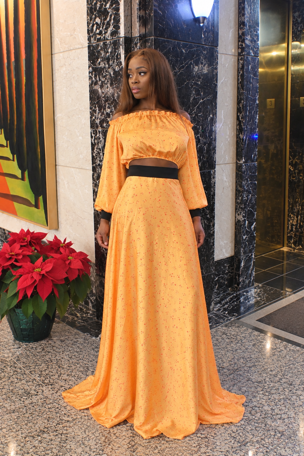 A woman wearing a bright orange two-piece maxi dress with a black waistband, standing in a lobby with marble columns and a potted poinsettia plant.