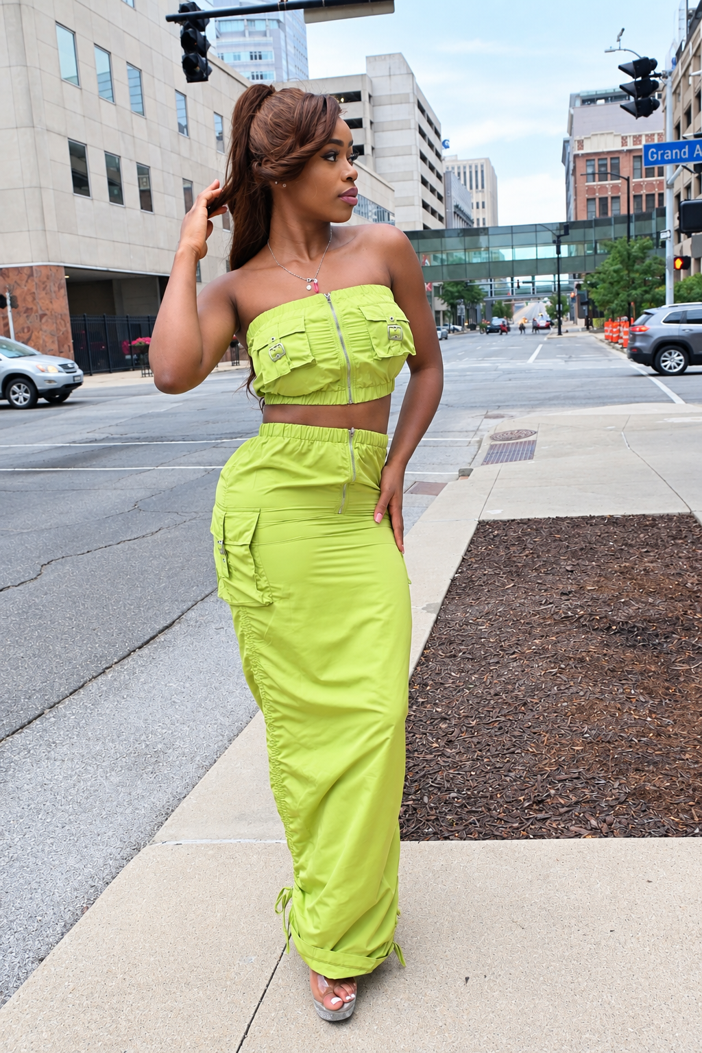 A woman stands on a city sidewalk wearing a bright lime green two-piece outfit consisting of a strapless crop top with flap pockets and a matching long skirt. The background shows a street scene with buildings, cars, and traffic lights.