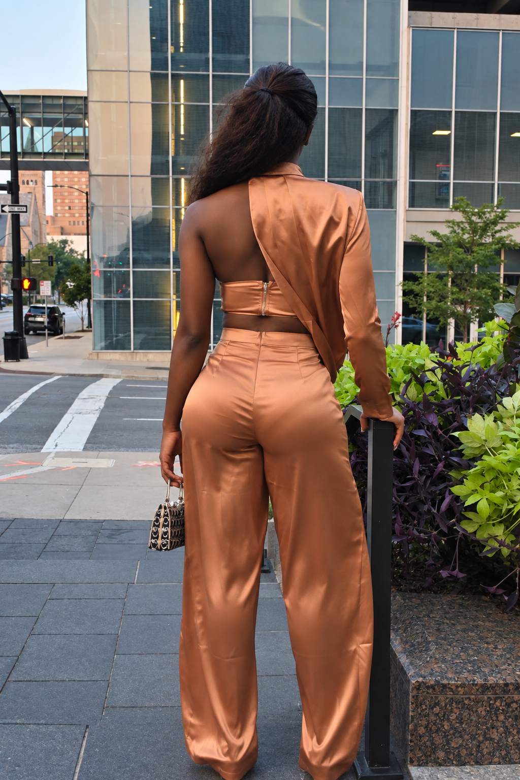 A woman stands on a city sidewalk facing away from the camera, wearing a matching satin three-piece outfit in a warm camel-brown color. The set includes an open-back long-sleeve crop top and high-waisted wide-leg pants.