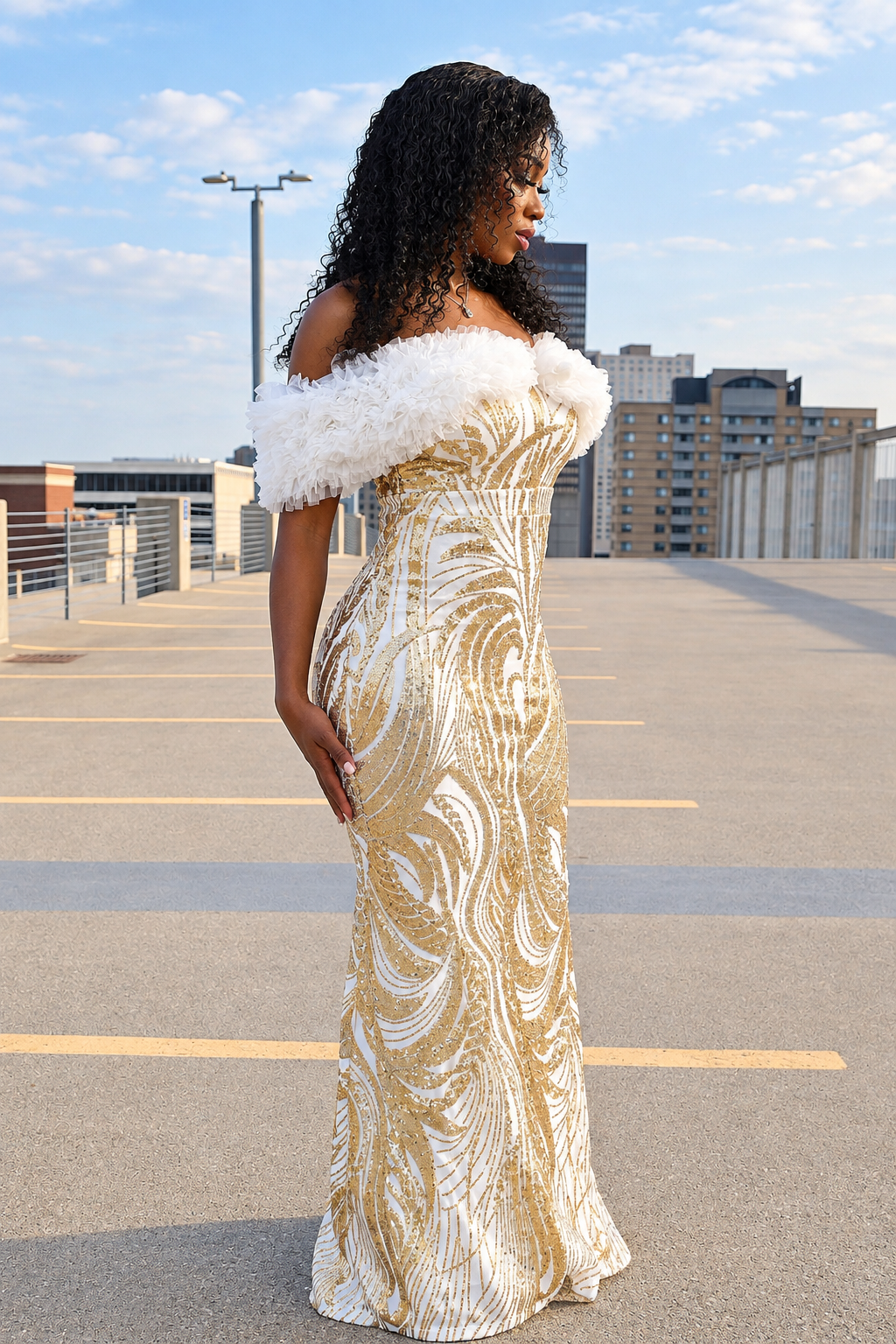 A woman stands on an outdoor rooftop wearing a floor-length off-the-shoulder gown with a white feathered shoulder detail and a gold and white swirling sequin pattern. The background shows a city skyline and blue sky.