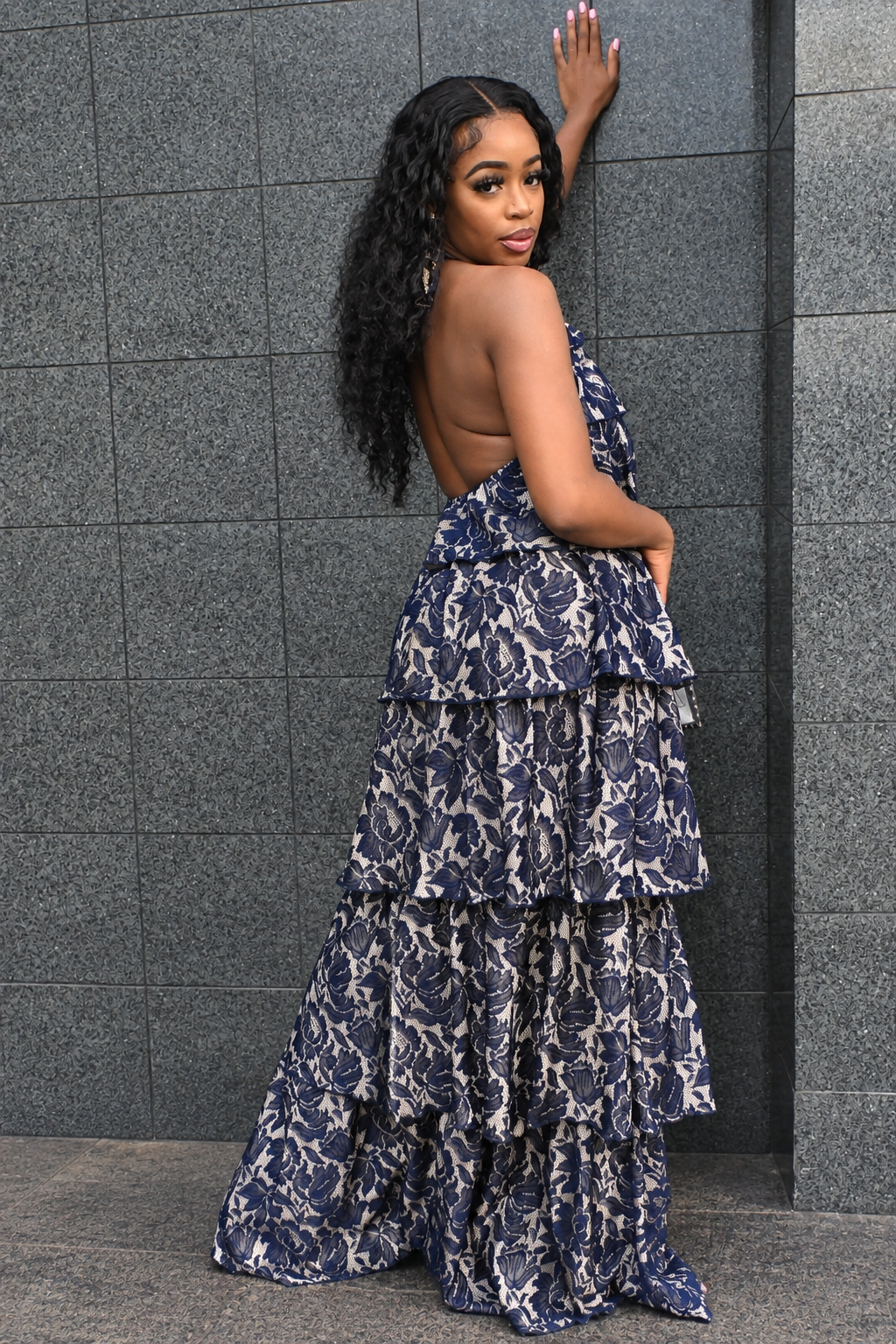 A woman wearing a navy and beige floral print tiered maxi dress with an open back, posing against a gray tiled wall.