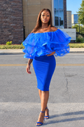 A woman modeling a bright royal blue off-the-shoulder dress with dramatic layered ruffle sleeves and a fitted knee-length skirt, paired with matching blue high-heeled sandals, standing outdoors on a paved street.