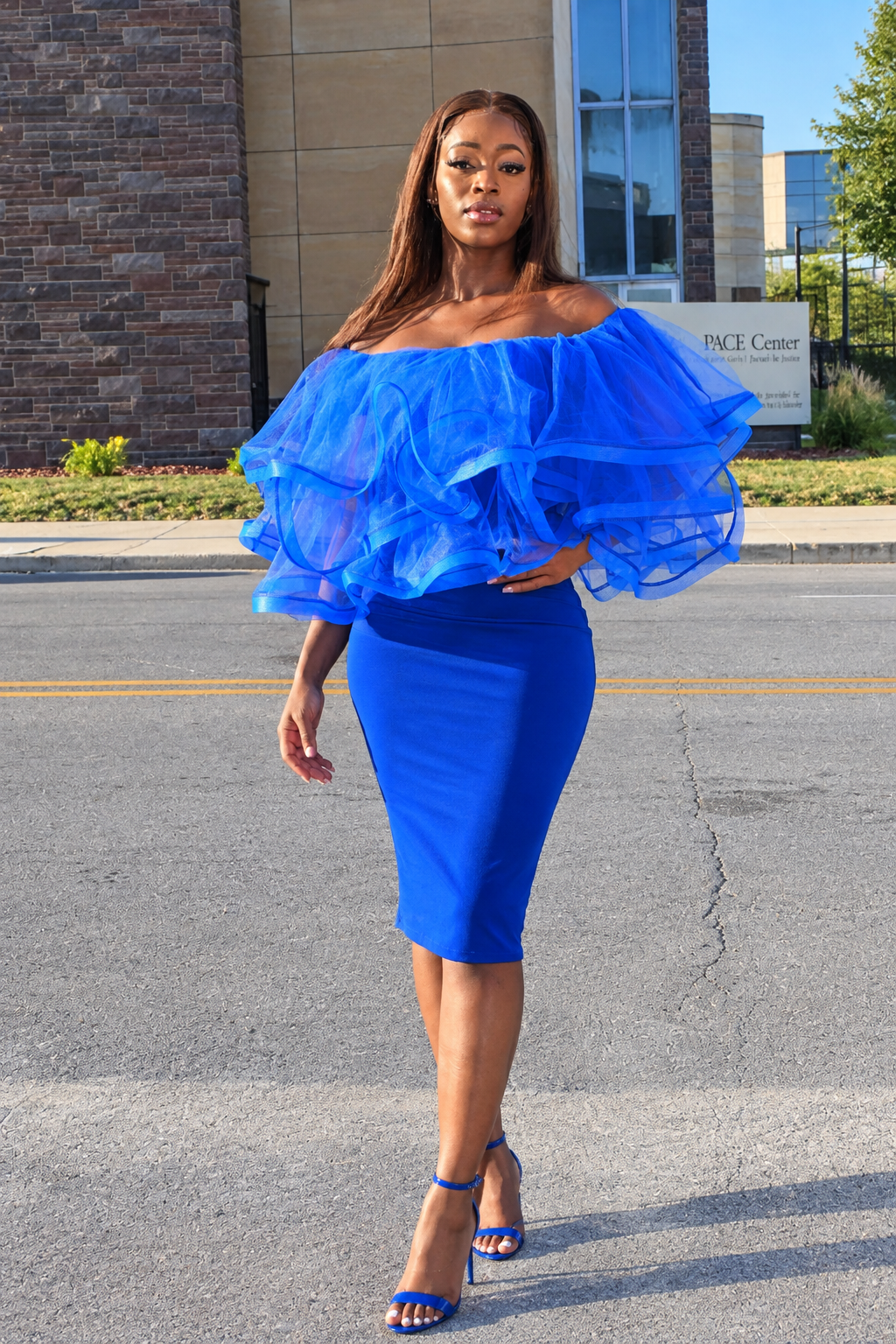 A woman modeling a bright royal blue off-the-shoulder dress with dramatic layered ruffle sleeves and a fitted knee-length skirt, paired with matching blue high-heeled sandals, standing outdoors on a paved street.