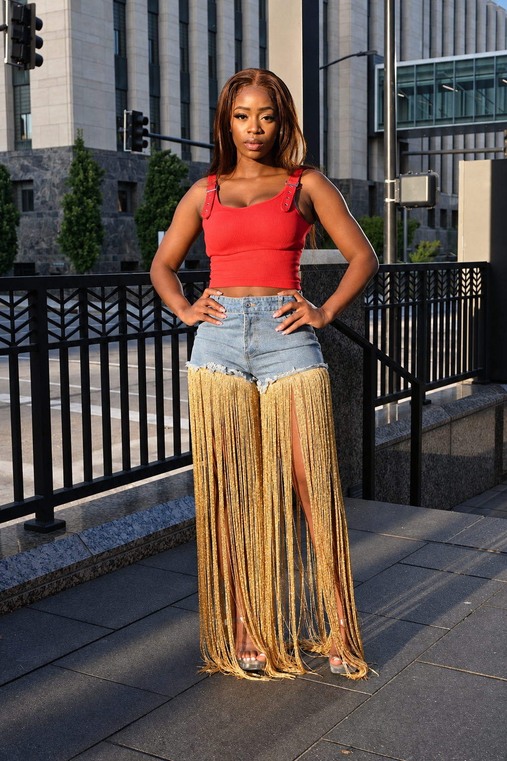 A woman stands outdoors on a city sidewalk wearing a bright red sleeveless crop top, light blue denim shorts, and a long fringe skirt in a warm metallic gold color. The background features a modern building, a black metal railing, and street elements.