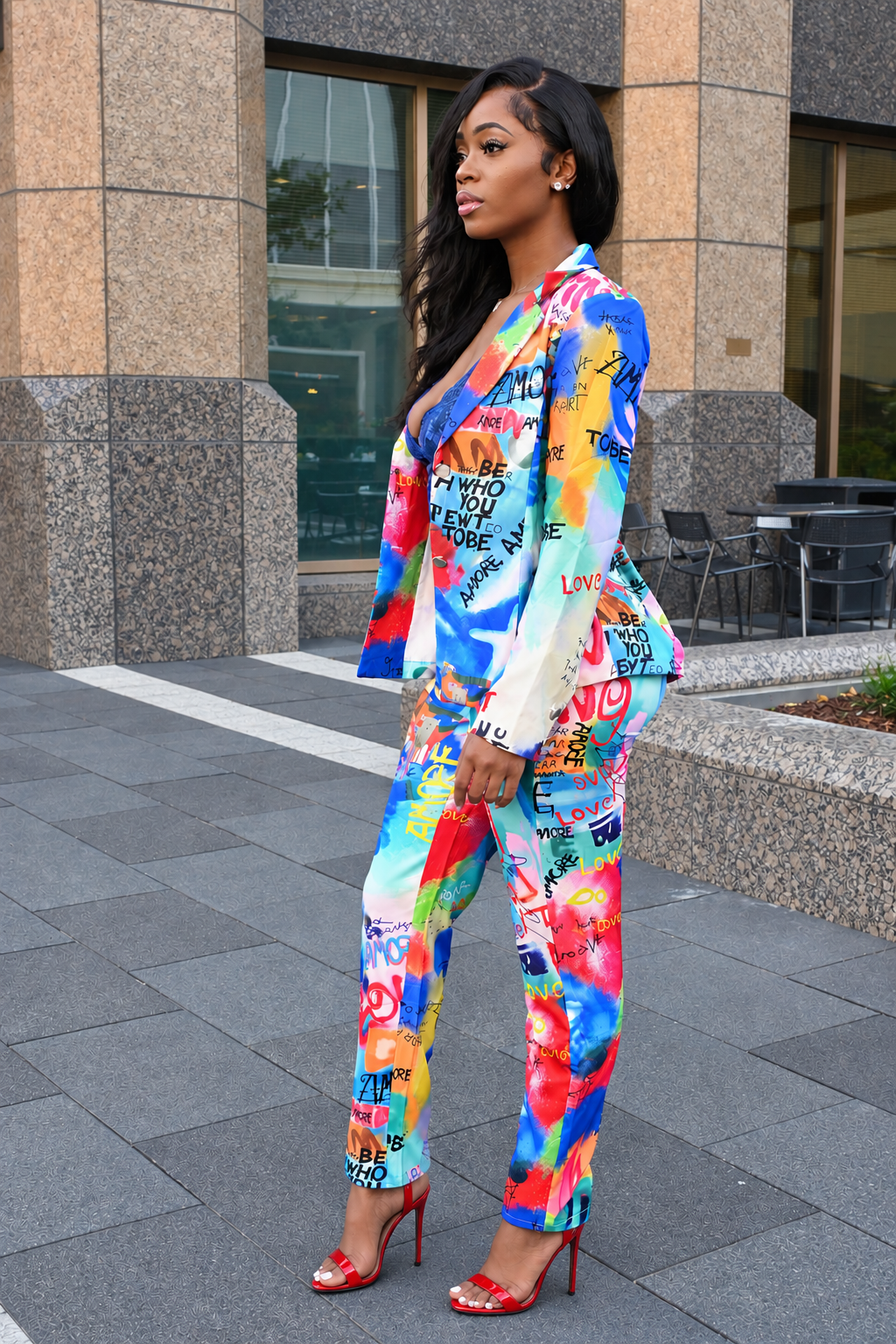 A woman stands outdoors in front of a modern building, wearing a bright multicolor graffiti-print blazer and matching pants with red high heels.