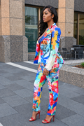 A woman stands outdoors in front of a modern building, wearing a bright multicolor graffiti-print blazer and matching pants with red high heels.