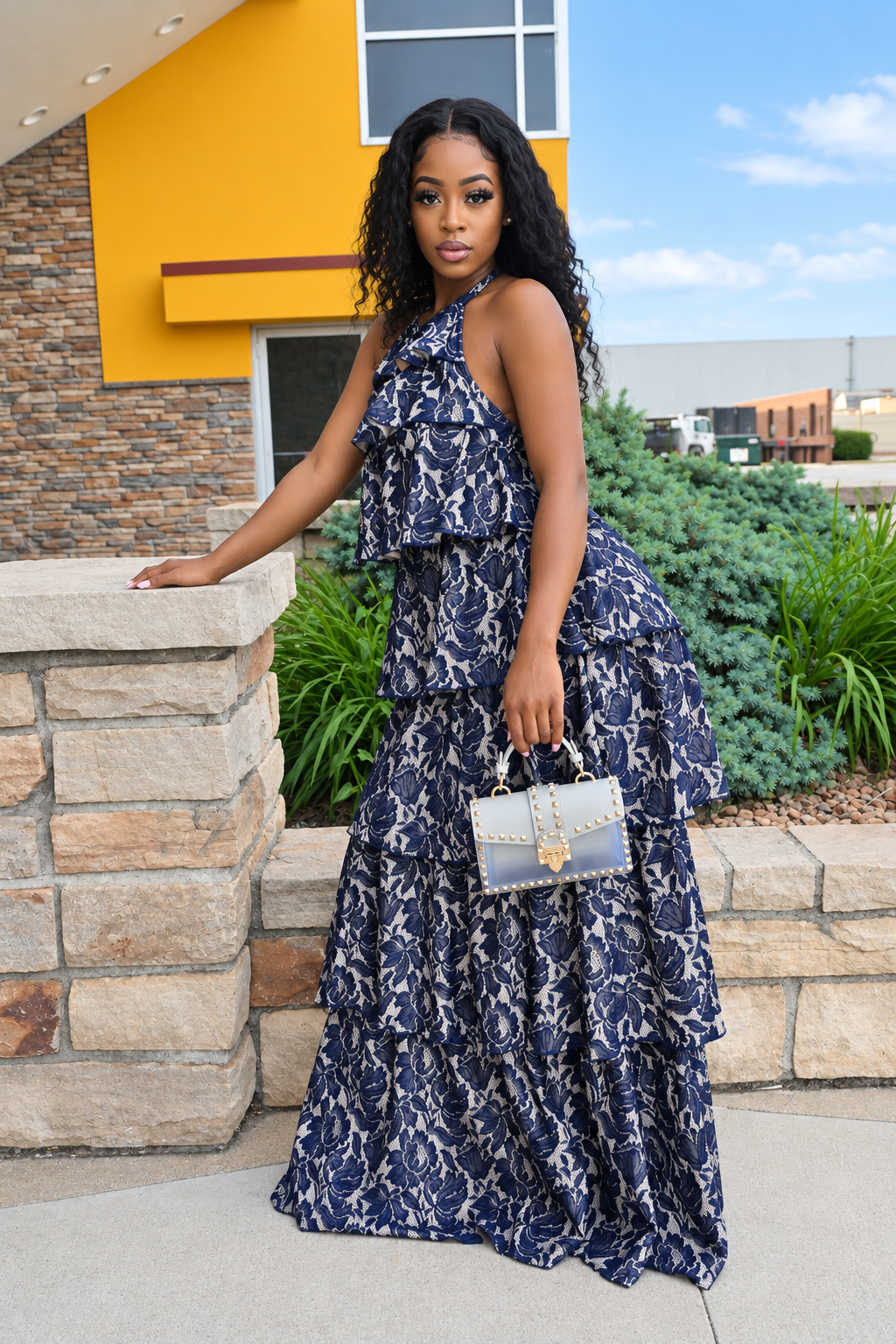 A woman stands outdoors wearing a navy and white snakeskin print halter-neck maxi dress with a tiered ruffle bodice and full skirt. She is holding a small silver and blue handbag with gold hardware, posing in front of a modern building with a yellow facade and stone walls.