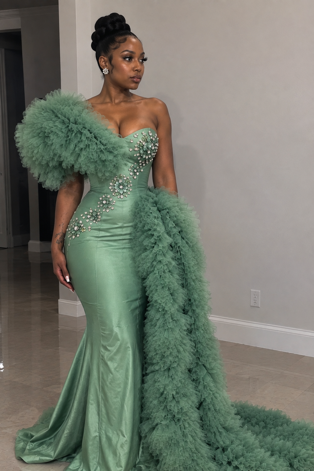 A woman models a strapless sage green gown with a sweetheart neckline, embellished beaded bodice, and a voluminous feathered overskirt and matching train in an indoor setting.