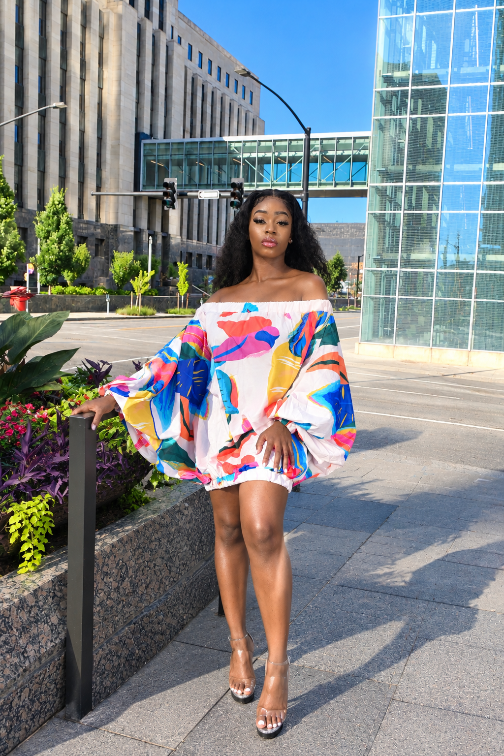 A woman stands on a city sidewalk wearing a colorful off-the-shoulder mini dress with dramatic balloon sleeves. The dress features a bold abstract print in blue, pink, yellow, orange, and green on a white base.
