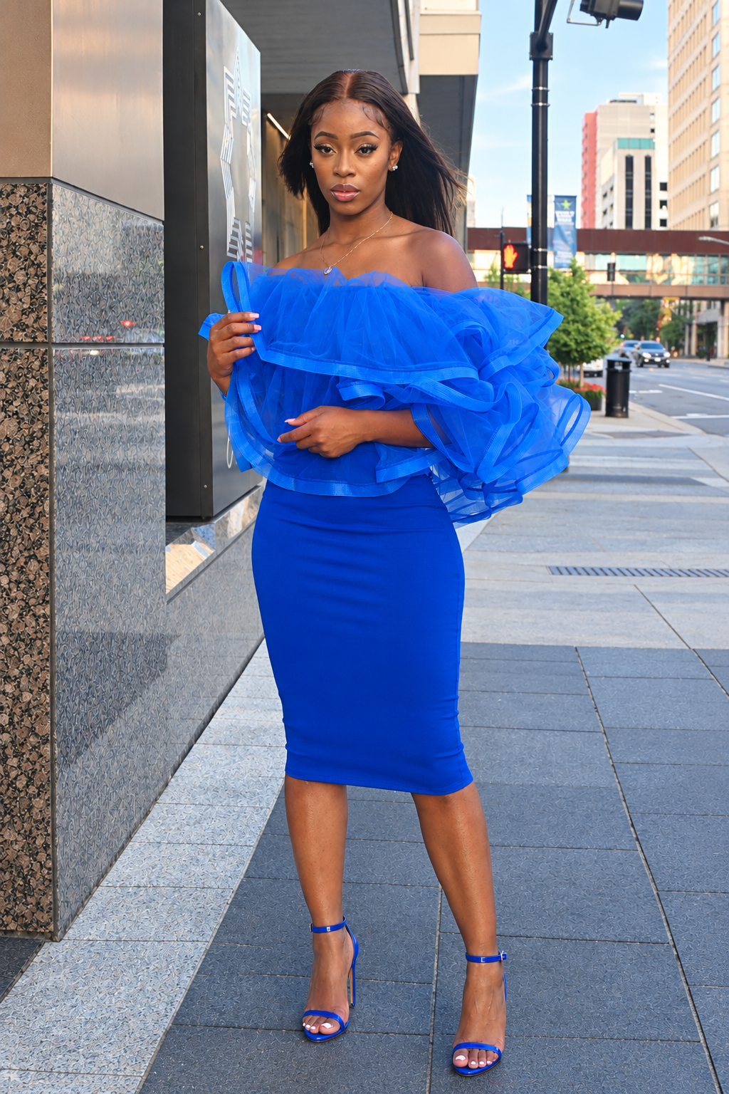 A woman stands on a city sidewalk wearing a bright royal blue off-the-shoulder ruffled top and matching fitted midi skirt, paired with blue high-heel sandals.