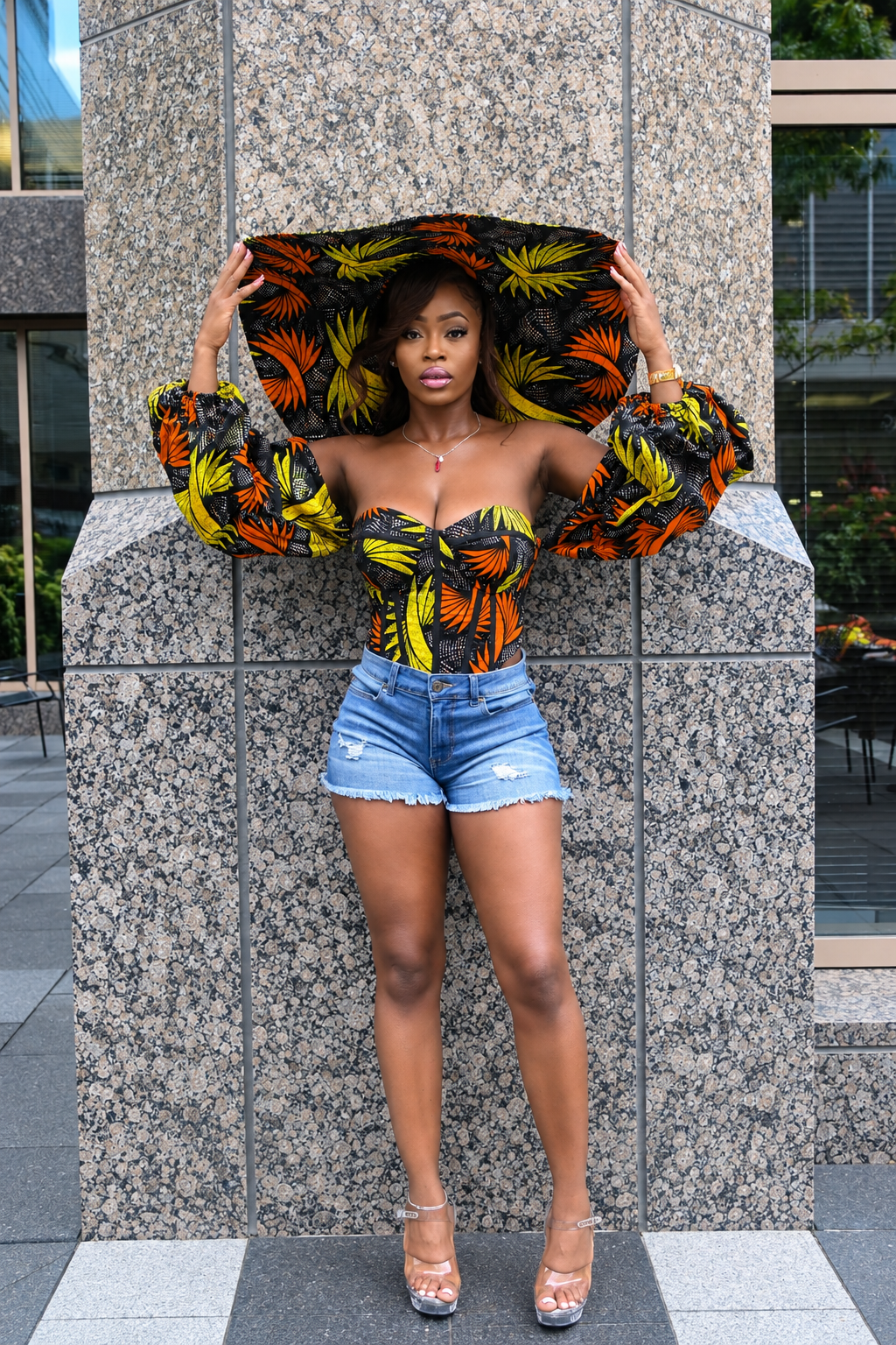 A woman stands outdoors in front of a large gray stone pillar, wearing a strapless fitted top with dramatic puffed sleeves in a bold tropical leaf print, blue denim shorts, and clear high heels. She is holding up a matching oversized hat or visor with the same print.