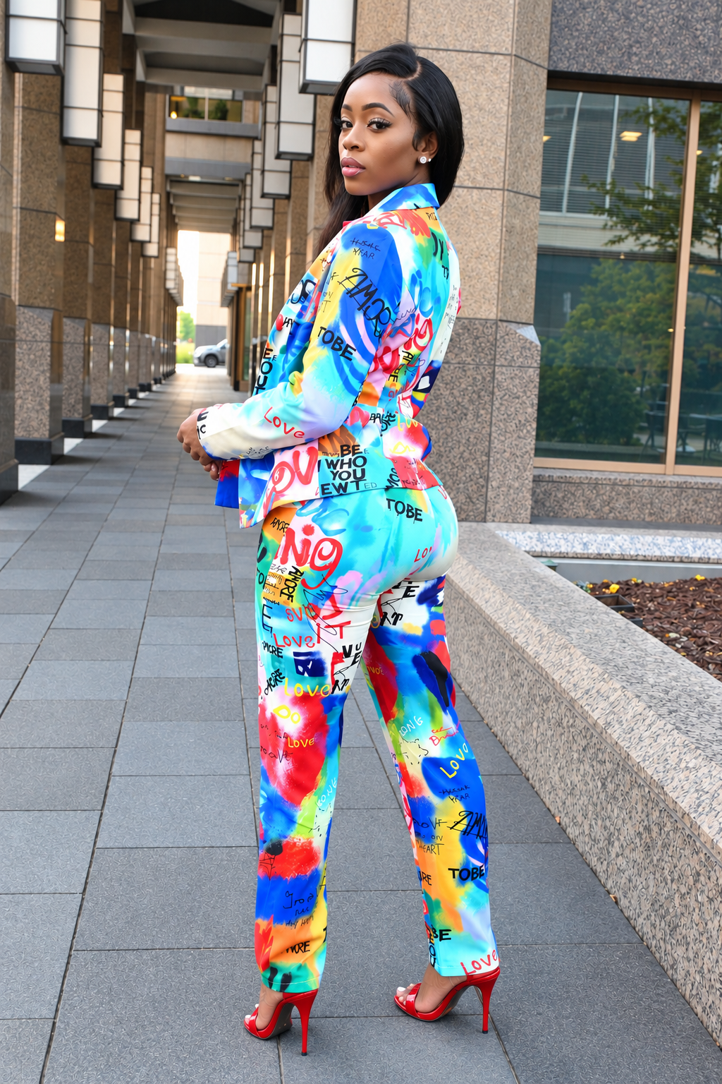A woman posing outdoors in a vibrant, multicolor graffiti-printed two-piece suit with red high heels, standing on a paved walkway next to a modern building.
