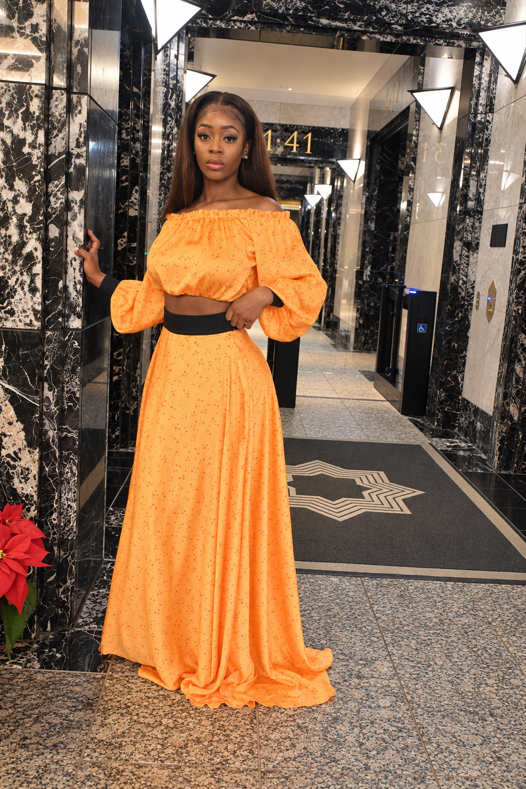 A woman stands in a hotel lobby wearing a bright orange off-the-shoulder maxi dress with a black waistband. The background features black and white marble columns and a patterned floor mat.