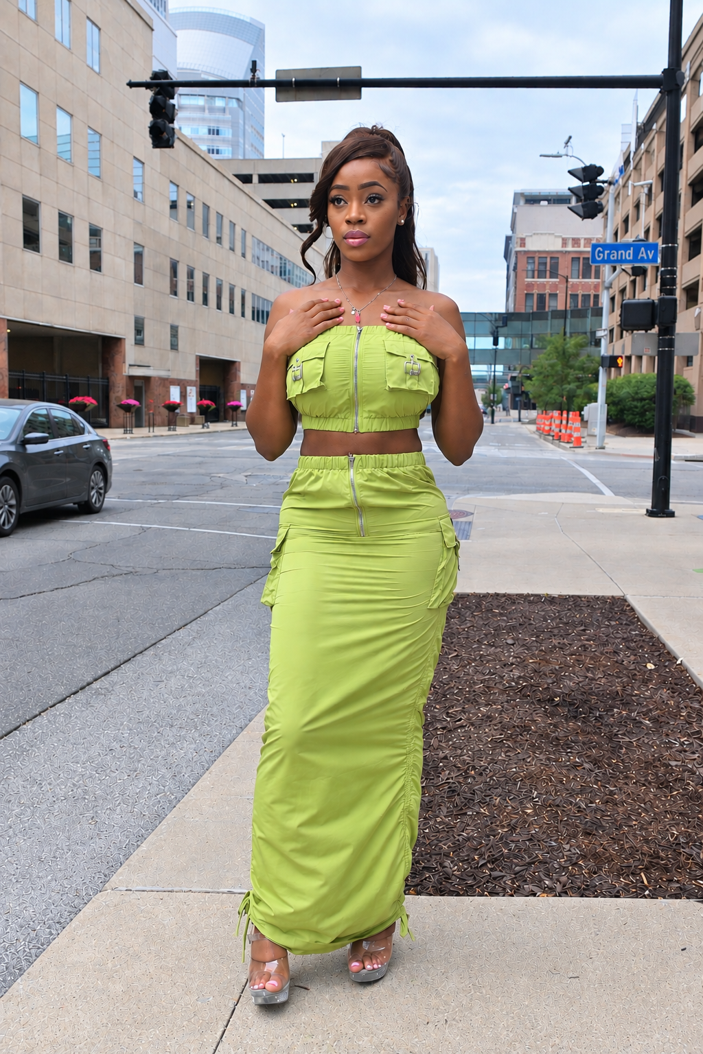A woman stands on a city sidewalk modeling a bright lime green two-piece outfit consisting of a strapless crop top with front pockets and a matching ruched maxi skirt.
