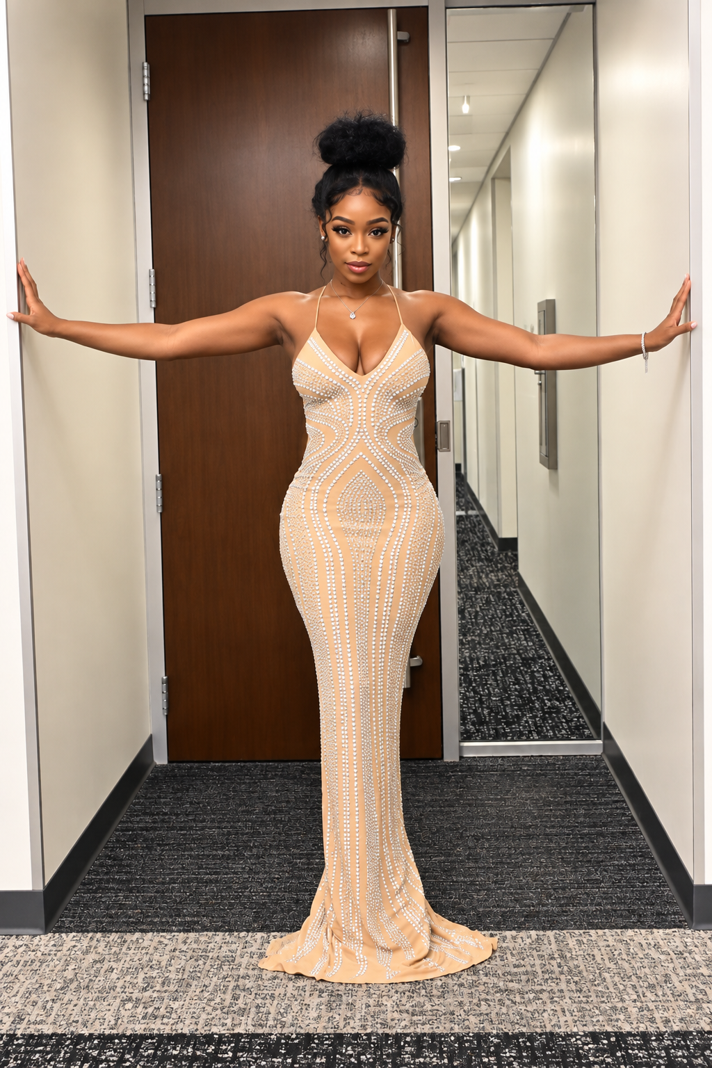A woman stands in a hallway wearing a fitted, floor-length beige gown with a geometric pattern of metallic gold and silver rhinestones. She has her hair in a high bun and is posing with her arms outstretched against the walls.