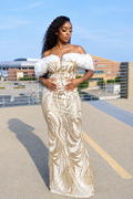 A woman wearing a floor-length off-the-shoulder gown with white ruffled puff sleeves and a metallic gold and white swirling pattern, standing outdoors on a sunny day.