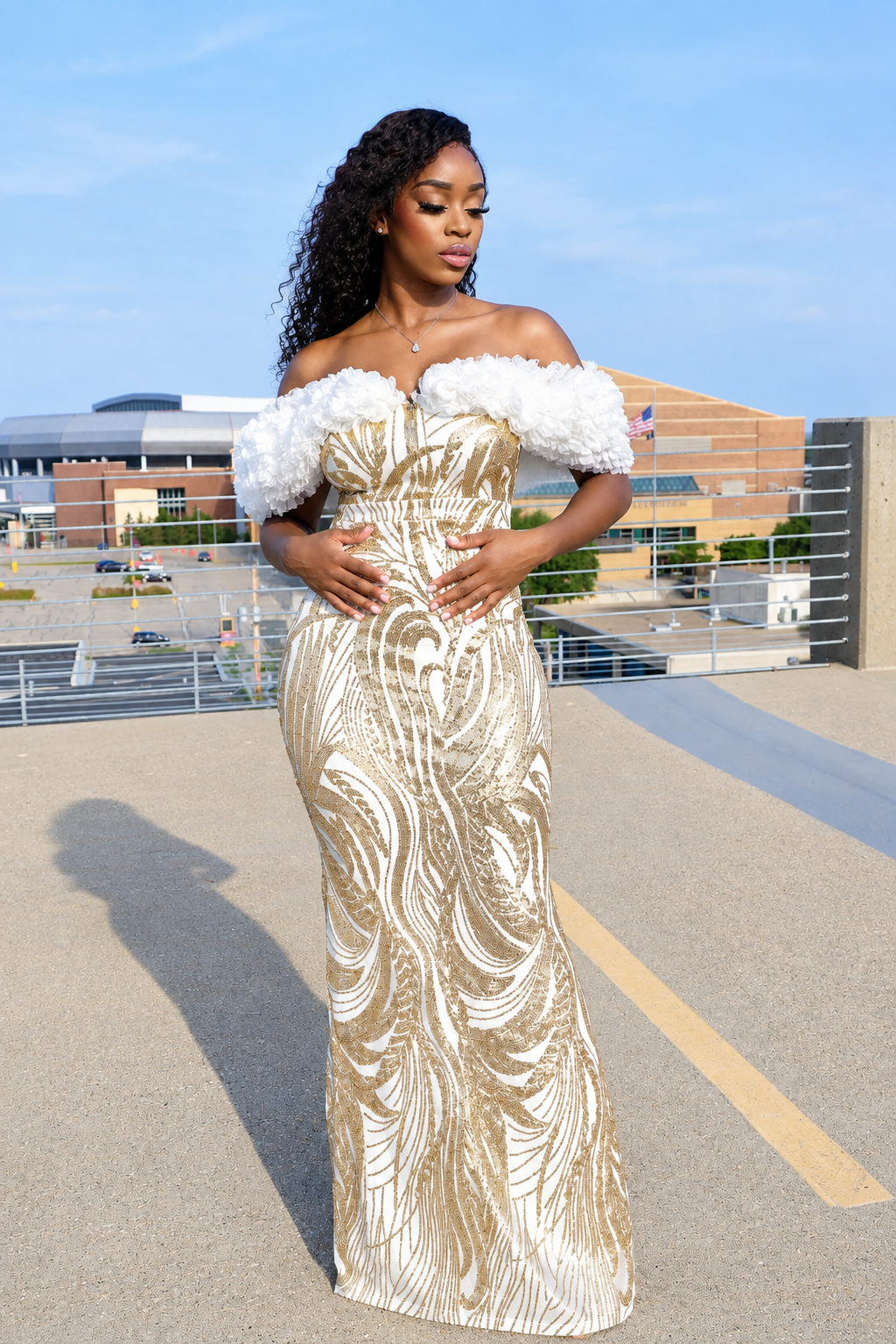 A woman wearing a floor-length off-the-shoulder gown with white ruffled puff sleeves and a metallic gold and white swirling pattern, standing outdoors on a sunny day.