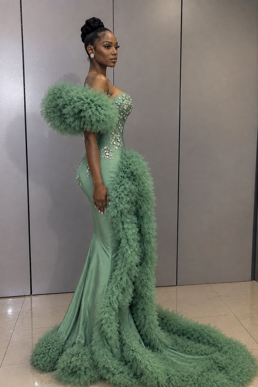 A woman modeling a floor-length sage green gown with dramatic feathered ruffle details, a structured sweetheart bodice with embellished crystal-like embroidery, and a long feather-trimmed train, posed against a gray paneled wall.