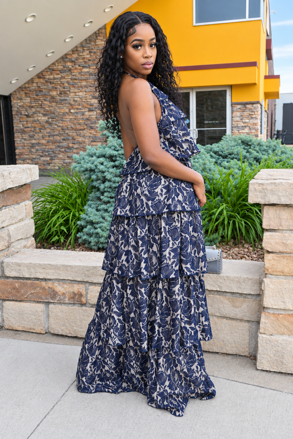 A woman wearing a navy blue and white floral print tiered maxi dress with an open back, standing outdoors in front of a modern building with a yellow facade and stone walls.