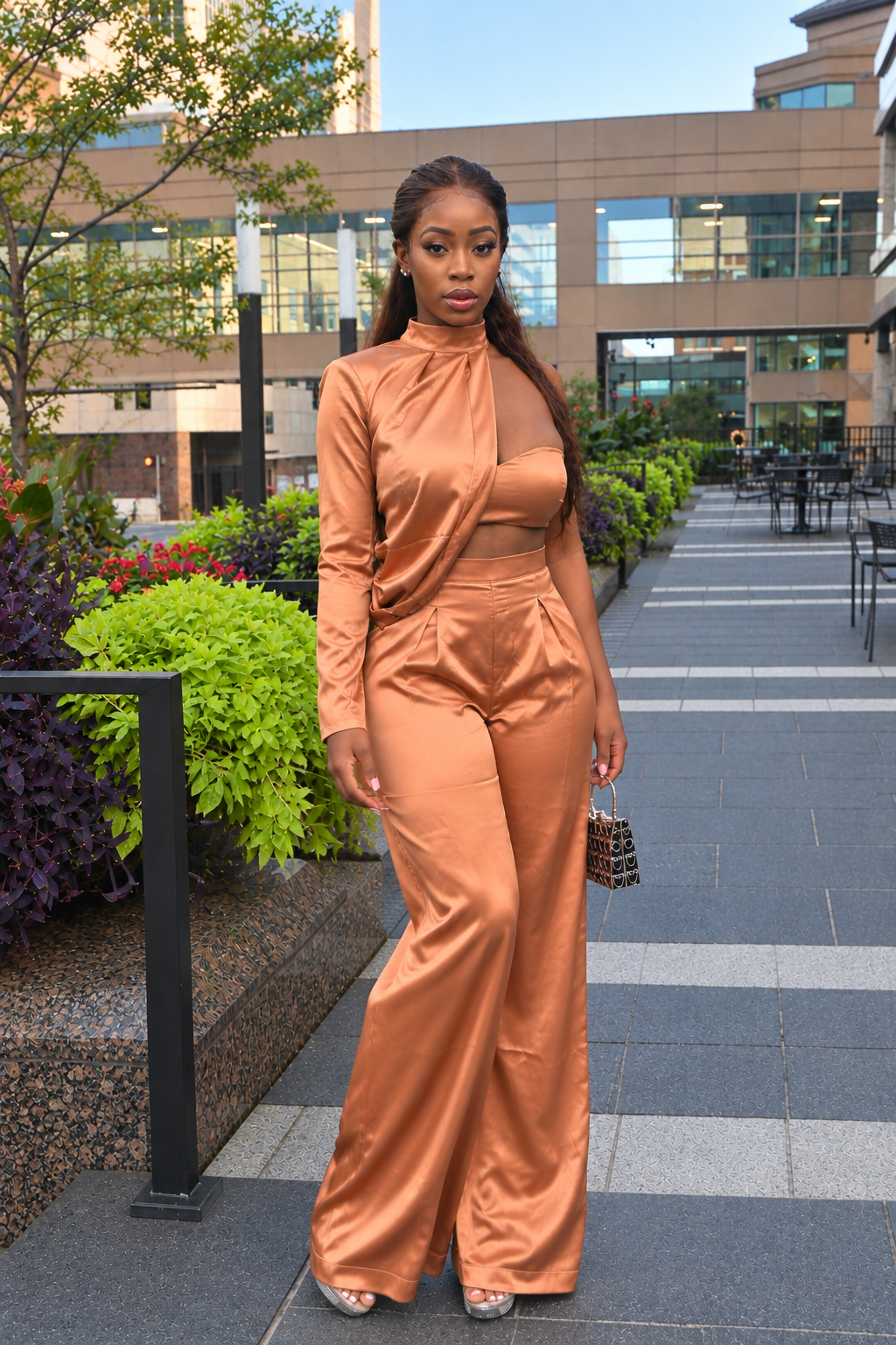 A woman stands outdoors on a paved walkway wearing a shiny copper-orange satin three-piece outfit with a high-neck long-sleeve crop top and matching wide-leg pants. She is holding a small patterned handbag, with a modern building and landscaped greenery in the background.
