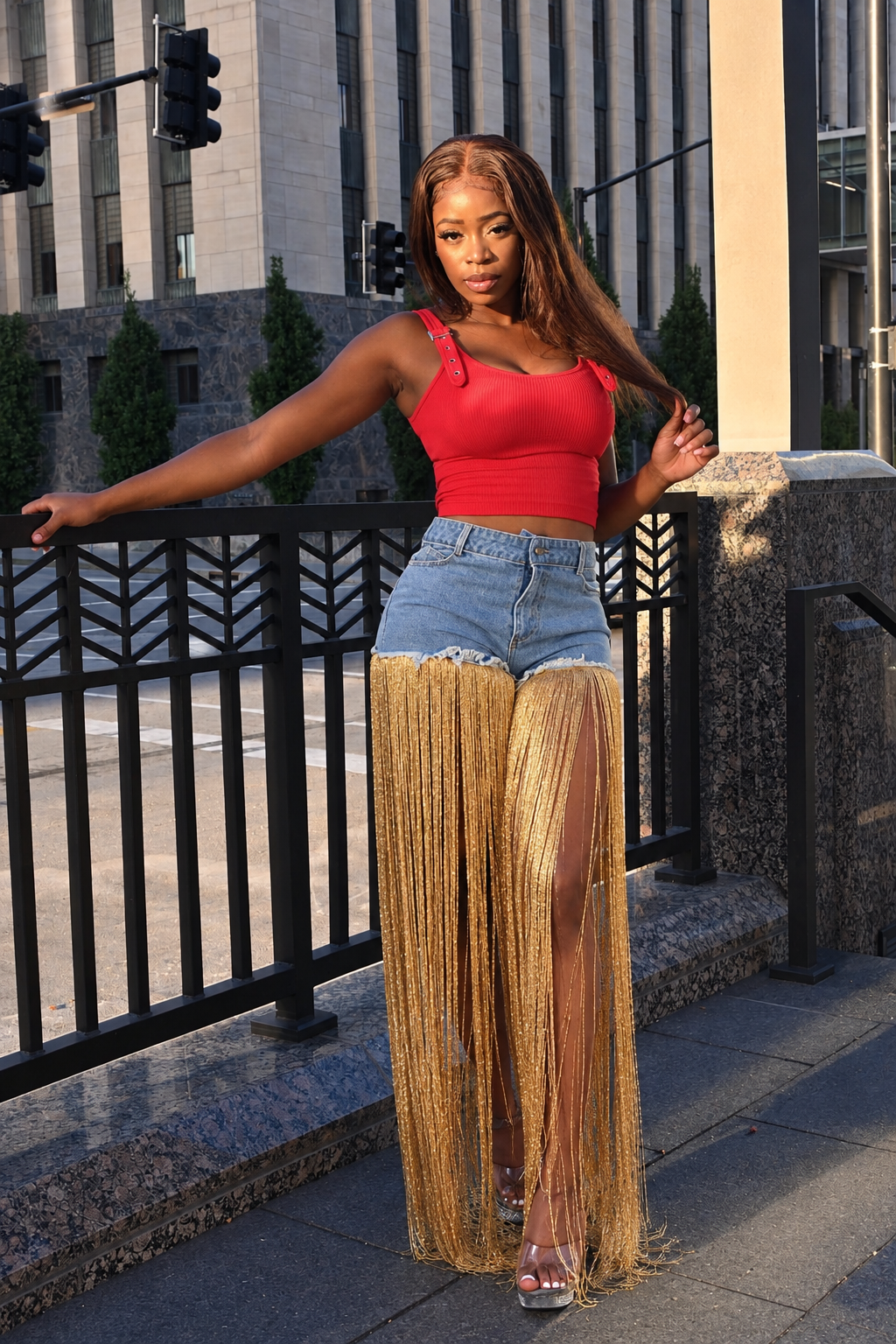 A woman posing outdoors on a city sidewalk next to a black metal railing, wearing a bright red ribbed tank top, light-wash denim shorts, and a long, shimmering gold fringe skirt.