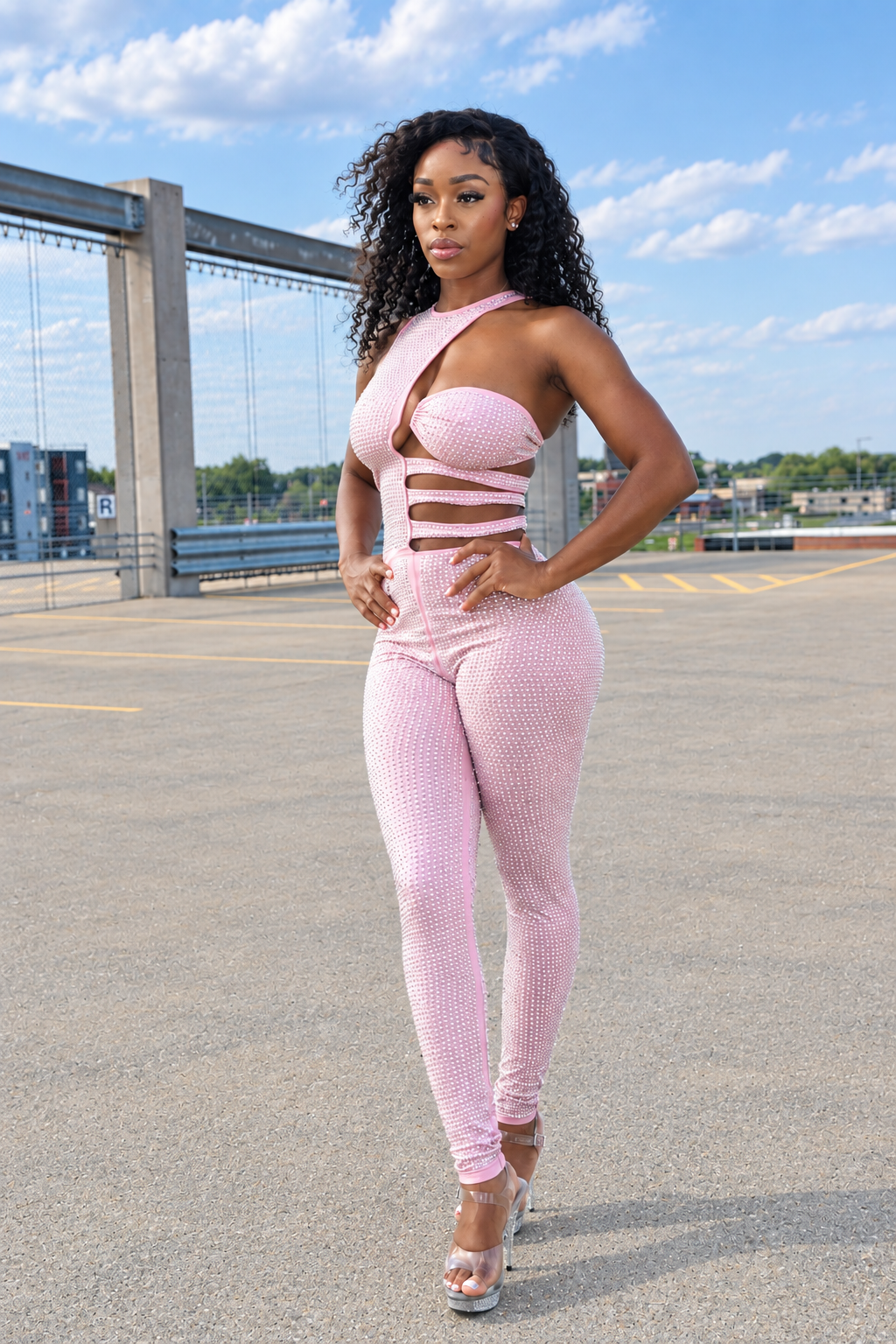 A woman stands outdoors in a parking lot wearing a light pink ribbed one-piece jumpsuit with a halter-style neckline, front cutouts, and matching leggings. She is wearing clear high-heeled sandals, with a bridge and blue sky in the background.