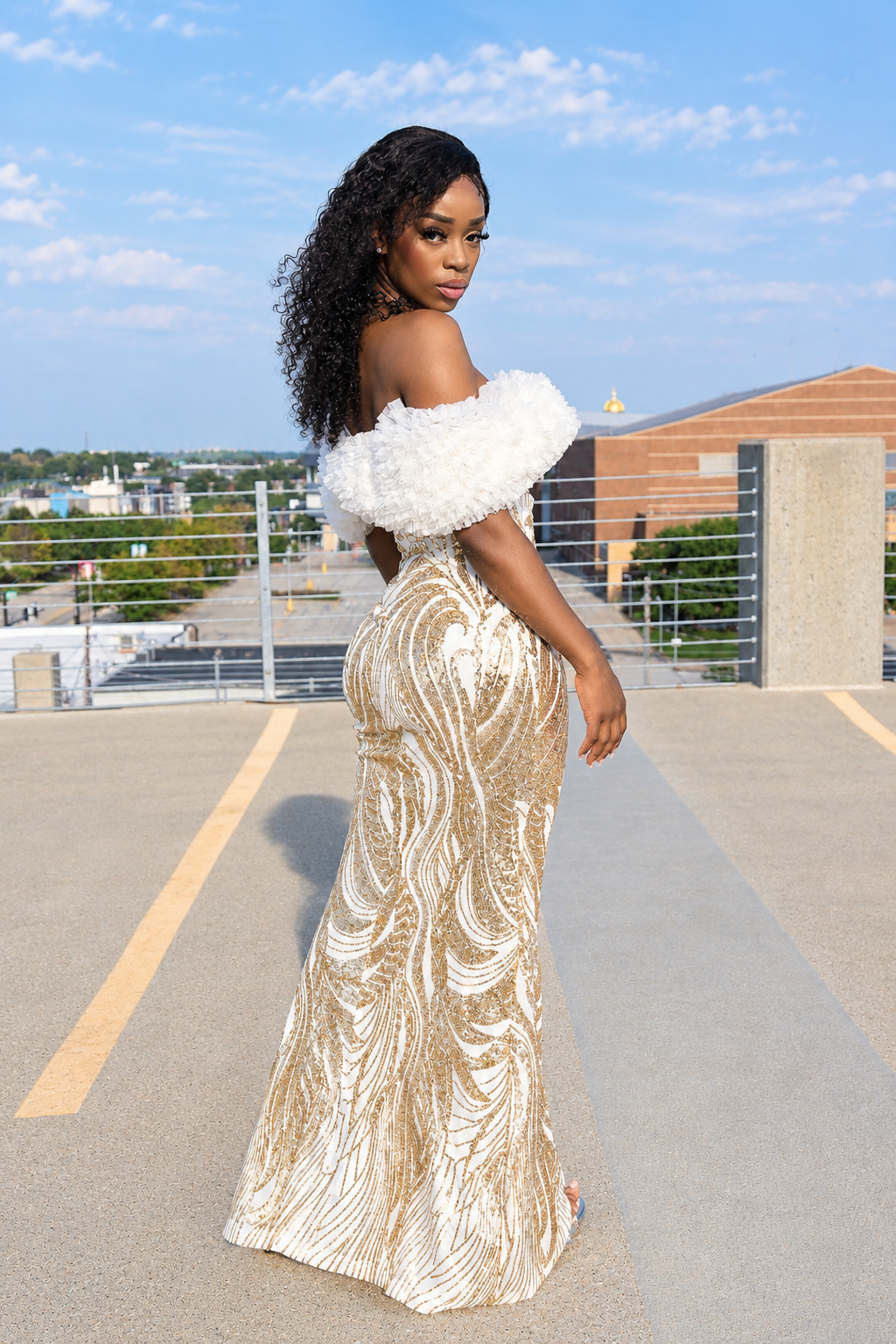 A woman wearing a floor-length off-the-shoulder gown with a white ruffled neckline and a gold sequined body, standing on an outdoor rooftop or elevated walkway under a blue sky.