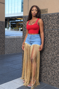 A woman stands outdoors against a speckled stone wall wearing a bright red ribbed tank top with decorative straps, light blue denim shorts, and a long beige fringe skirt overlay. She is wearing clear high-heel sandals.